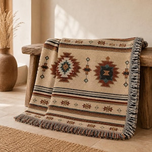 May include: A woven throw blanket featuring a beige base with repeating geometric patterns in shades of rust, navy blue, and dark brown. The blanket has horizontal striped borders and decorative fringe along the edges, displaying a textured fabric construction.