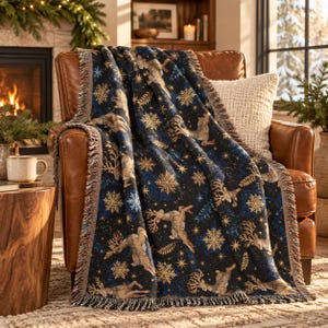 May include: A woven throw blanket featuring a dark blue background with a repeating pattern of tan reindeer, gold snowflakes, and small stars. The blanket has a fringed edge and is draped over a brown leather armchair with a textured white pillow.