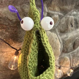 May include: A lime green crocheted creature with a large open mouth, two white spherical eyes with dark pupils, and purple pipe cleaner antennae. The item features a hanging loop at the top and a textured, yarn-based construction.