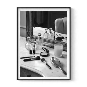 Op de afbeelding: Een zwart-wit ingelijste print met een kaptafel met spiegel, glazen parfumflesjes, lippenstiften, make-upkwasten, een poederdoosje en een haarborstel. De scène toont een verzameling schoonheidsaccessoires gerangschikt op een marmeren oppervlak.