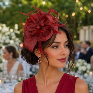 Puede incluir: Un tocado fascinator de color rojo intenso con una base de tela tejida texturizada, adornado con grandes bucles estructurados y plumas finas y delicadas. El accesorio se combina con un vestido rojo a juego y pendientes de piedra roja en forma de gota.
