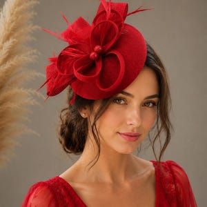 Könnte beinhalten: Ein leuchtend roter Fascinator mit einer kreisförmigen Basis, strukturierten Schlaufen aus versteiftem Stoff und zarten, aufrechten Federn. Zwei kleine, runde, passende rote Perlen befinden sich in der Mitte der dekorativen Schlaufenanordnung.