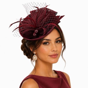 Pode incluir: Um fascinator em tom bordô profundo com uma base estruturada, laços de tecido complexos e penas delicadas. A peça é acentuada com um véu de rede fina e dois pequenos ornamentos esféricos texturizados num tom vermelho escuro a condizer.
