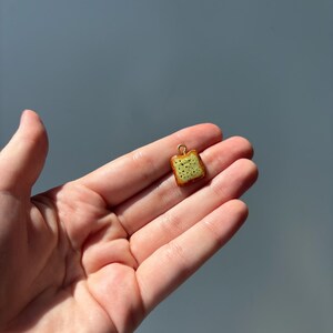 May include: A small, square-shaped charm shaped like a slice of toast with a golden-brown crust and a light yellow, speckled surface. The charm features a small, circular gold-toned metal loop at the top for attachment to a chain or bracelet.