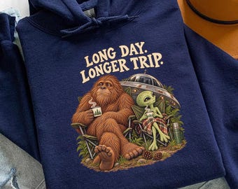 Könnte beinhalten: Ein marineblauer Kapuzenpullover mit einem Grafikdruck eines braunen Bigfoot und eines grünen Außerirdischen, die mit Tassen in Campingstühlen sitzen. Der Text über der Illustration lautet LONG DAY. LONGER TRIP. Der Hoodie hat lange Ärmel.