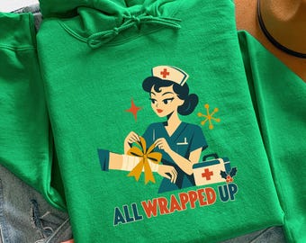 Könnte beinhalten: Ein hellgrüner Kapuzenpullover mit einer Grafik einer Krankenschwester in Haube, die einen Verband mit einer gelben Schleife um einen Arm wickelt. Neben der Krankenschwester steht ein Erste-Hilfe-Kasten. Der Text "ALL WRAPPED UP" steht in Blau und Orange.