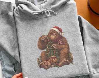 Könnte beinhalten: Ein sportgrauer Kapuzenpullover mit einer Grafik eines braunen Orang-Utans, der eine rot-weiße Weihnachtsmütze und einen gemusterten Schal trägt. Der Orang-Utan schmückt einen kleinen Weihnachtsbaum mit Kugeln und Geschenken.