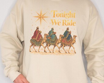 Könnte beinhalten: Ein beigefarbenes Sweatshirt mit Rundhalsausschnitt und einem Grafikdruck von drei Figuren auf Kamelen in einer Wüstenlandschaft. Über den Figuren befindet sich ein goldener Stern und der Text Tonight We Ride in Gold, Rot und Blau.