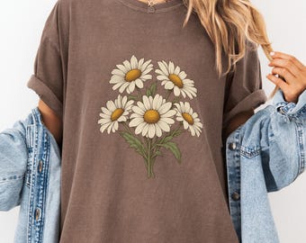 Könnte beinhalten: Ein espressofarbenes Kurzarm-T-Shirt mit einer zentralen Grafik eines Blumenstraußes aus weißen Gänseblümchen mit gelben Mitten und grünen Stielen. Das T-Shirt hat einen Rundhalsausschnitt und wird unter einem hellblauen Jeanshemd getragen.