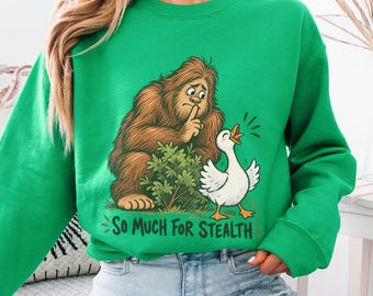 Könnte beinhalten: Ein hellgrünes Sweatshirt mit Rundhalsausschnitt und einer grafischen Illustration eines braunen Bigfoot, der sich hinter einem grünen Busch versteckt und einer weißen Gans ein Schweigezeichen gibt. Der Text lautet So Much For Stealth.