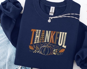 Könnte beinhalten: Ein marineblaues Sweatshirt mit Rundhalsausschnitt und dem Schriftzug THANKFUL in einer Mischung aus karierten und einfarbigen Buchstaben in Erdtönen. Unter dem Text befindet sich eine Kürbiszeichnung, umgeben von herbstlichen Blättern.