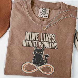 May include: An espresso-colored t-shirt featuring a graphic of a black cat sitting on an infinity symbol. The text above the cat reads NINE LIVES. INFINITE PROBLEMS. The shirt has a crew neckline and a Comfort Colors brand label inside the collar.