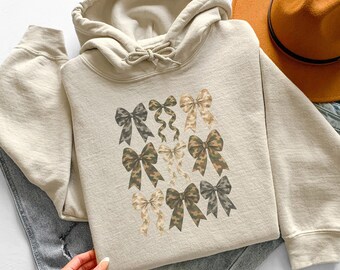 Könnte beinhalten: Ein beigefarbener Kapuzenpullover mit einem Grafikdruck von neun Schleifen in verschiedenen Braun-, Beige- und Olivgrüntönen mit Tarnmustern. Der Hoodie verfügt über eine passende Kordel am Ausschnitt und lange Ärmel, kombiniert mit einem braunen Filzhut.
