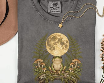 Könnte beinhalten: Ein anthrazitfarbenes T-Shirt mit Rundhalsausschnitt und einem Grafikdruck eines Frosches, der auf einem moosigen Baumstamm unter einem Vollmond sitzt, umgeben von Farnen und Pilzen. Das T-Shirt hat ein Comfort Colors Etikett am Hals.