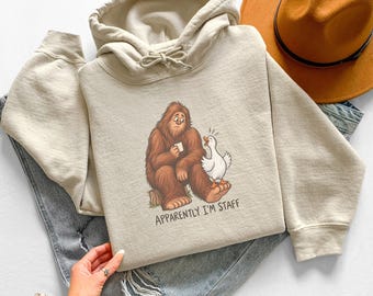 Könnte beinhalten: Ein beigefarbener Kapuzenpullover mit einer Grafik eines braunen Bigfoot, der eine Tasse hält, und einer weißen Gans, die auf seinem Fuß steht. Der Text unter der Abbildung lautet: "APPARENTLY I'M STAFF". Der Hoodie hat eine Kapuze mit Kordelzug.