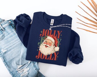 Könnte beinhalten: Ein marineblaues Sweatshirt mit Rundhalsausschnitt und einer Grafik eines zwinkernden Weihnachtsmanns mit weißem Bart und roter Mütze. Der Schriftzug JOLLY ist in Rot darüber und darunter gedruckt, mit dem Wort JOY in Grün dahinter.