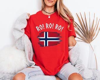 May include: A bright red short-sleeved t-shirt featuring the text RO! RO! RO! in white capital letters above a graphic of the Norwegian flag with a distressed, brushstroke-style border. The shirt has a crew neckline and a relaxed fit.