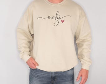 Könnte beinhalten: Ein beigefarbenes Sweatshirt mit Rundhalsausschnitt, auf dessen Brust das Wort mahj in schwarzer Schreibschrift steht, ergänzt durch ein kleines rosa Herzsymbol rechts neben dem Text. Das Kleidungsstück hat lange Ärmel und einen gerippten Kragen.