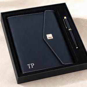 Könnte beinhalten: Ein schwarzes Notizbuch mit weißen Nähten und den in Weiß geprägten Initialen TP, ausgestattet mit einer dreieckigen Klappe und einem goldenen Metallverschluss, in einer schwarzen Geschenkbox mit passendem Kugelschreiber.