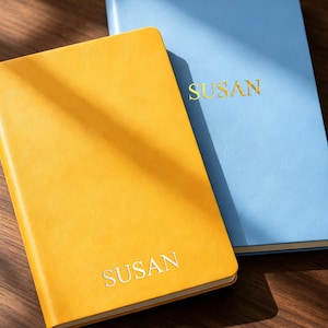 Op de afbeelding: Twee rechthoekige notitieboeken met gladde kaften, één in felgeel en één in lichtblauw. Op de voorkant van elk notitieboek staat de naam SUSAN in een strak, schreefloos lettertype gedrukt. Het gele notitieboek heeft zilveren letters en het blauwe gouden letters.
