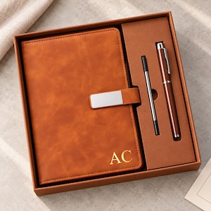 Könnte beinhalten: Ein hellbraunes Notizbuch mit den goldenen Buchstaben AC, einem silbernen Metallverschluss sowie einem passenden Stift mit silbernem Clip und einer separaten Tintenmine, präsentiert in einer rechteckigen Geschenkbox mit Fächern.