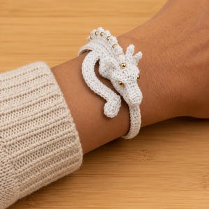 May include: A white crocheted dragon bracelet featuring a long, winding body that wraps around the wrist. The dragon has a textured, stitched surface with small, shiny gold-colored beads embedded along its spine and as eyes on its head.