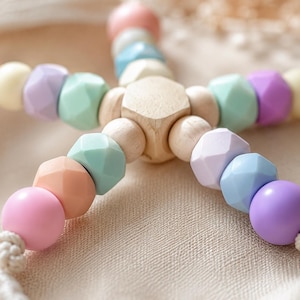 May include: A star-shaped toy constructed from a central wooden hexagon connected to five radiating strands of multi-colored beads. The beads feature a mix of smooth round and faceted geometric shapes in pastel shades of pink, peach, yellow, mint, and purple.