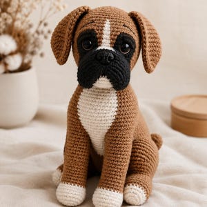 Pode incluir: Um bicho de pelúcia feito em crochê com a forma de um cão boxer. O brinquedo apresenta um corpo castanho com peito branco, patas brancas e um focinho preto. Possui olhos grandes, escuros e redondos e orelhas caídas feitas com fios.