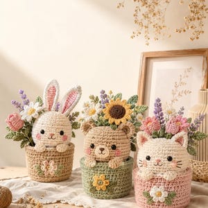 May include: Three crocheted animal figures in baskets: a white rabbit in a tan basket, a tan bear in a green basket, and a white cat in a pink basket. Each basket is decorated with a crocheted flower and filled with crocheted flowers and greenery.