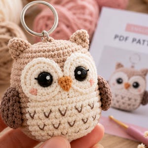 Pode incluir: Um porta-chaves de coruja em croché com cabeça castanho-clara, asas castanho-escuras e barriga creme com bordados em forma de V. A coruja tem grandes olhos pretos de plástico, um pequeno bico laranja e detalhes de blush rosa nas bochechas.