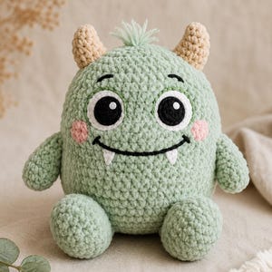 May include: A light green crocheted monster plush toy with two tan horns, a small tuft of hair on top, large black and white eyes, pink circular cheeks, and a black stitched mouth with two small white fangs. The creature has a round body.