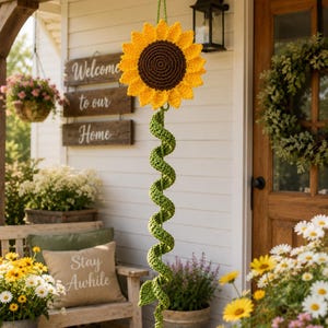 May include: A hanging crochet sunflower decoration featuring a large yellow flower head with a dark brown center, attached to a long, spiraling green stem made of yarn. The sunflower is suspended by a green cord and includes a small green leaf.