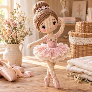 Puede incluir: Una muñeca bailarina de ganchillo con cabello de hilo marrón claro recogido en un moño, que lleva un leotardo rosa y un tutú de tul en capas. La muñeca presenta detalles faciales bordados, mejillas rosadas y pequeñas flores blancas decorativas.