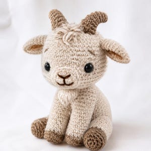May include: A crocheted stuffed goat toy in a sitting position, featuring a light beige body, darker brown curved horns, and brown hooves. The goat has large black safety eyes, a stitched nose and mouth, and a small tuft of yarn on its chin.