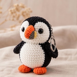 May include: A crochet puffin keychain featuring a black and white body, round black eyes, and a multi-colored beak with orange, yellow, and grey sections. The bird has bright orange feet and a silver metal keyring attached to its side.