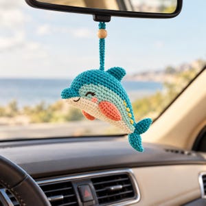 Könnte beinhalten: Ein gehäkelter Delfin als Hängedekoration mit einem Körper in Türkis und Hellblau, pfirsichfarbenen Flossen und einer kleinen Holzperle an der Aufhängung. Der Delfin hat gestickte geschlossene Augen und einen lächelnden Mund.