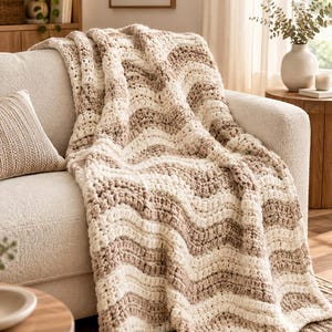 May include: A chunky knit throw blanket featuring a wavy chevron pattern in alternating stripes of cream and light beige. The blanket is crafted from thick, soft yarn with a visible textured stitch, draped over the arm and seat of a sofa.