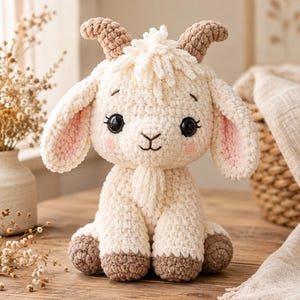 May include: A cream-coloured crochet goat plush toy with brown horns and hooves. The stuffed animal features large black safety eyes, a small tuft of hair on its head, a goatee, and pink blush accents on its cheeks. The texture is soft and fuzzy.