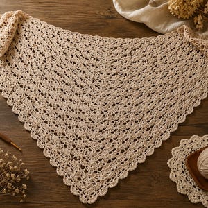 May include: A triangular beige crochet shawl featuring an intricate repeating shell stitch pattern with a scalloped edge. The shawl is accompanied by a wooden crochet hook, a ball of matching yarn on a small coaster, and dried flowers.