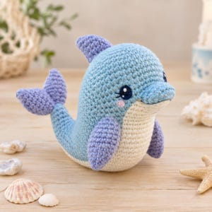 Op de afbeelding: Een gehaakt dolfijn knuffeltje met een lichtblauw lichaam, een crèmekleurige buik en paarse vinnen en staart. De dolfijn heeft grote, donkerblauwe veiligheidsoogjes met witte accenten en kleine roze geborduurde wangen op het gezicht.