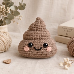 May include: A brown crocheted plush toy shaped like a swirl with a smiling face, featuring two black plastic eyes, a stitched black mouth, and two small pink circular cheeks. The yarn texture is visible throughout the entire surface of the object.