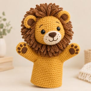 Puede incluir: Una marioneta de mano de ganchillo con forma de león, cuerpo amarillo dorado y una melena gruesa de lana marrón oscuro. El león tiene un hocico color crema, nariz marrón oscuro, ojos negros y almohadillas marrones en sus dos brazos extendidos.