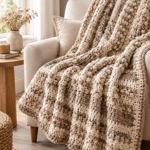 May include: A crochet throw blanket featuring a textured pattern of alternating vertical stripes in shades of cream, beige, and light brown. The design incorporates raised bobble stitches and smooth knit-like sections, creating a tactile surface.