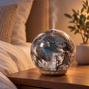 May include: A spherical glass essential oil diffuser with a dark, marbled, and textured pattern in shades of blue, grey, and black. The device features a light-coloured wooden base with three small control icons and emits a thin stream of white mist.