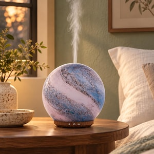 May include: A spherical essential oil diffuser with a marbled blue, white, and purple glass exterior. The device features a dark wood-toned base with three small control buttons and emits a steady vertical stream of mist from the top centre.