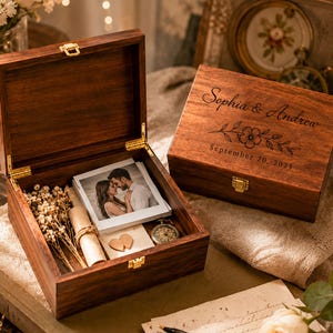 May include: Two dark brown wooden keepsake boxes with gold-tone latches. One box is open, revealing a stack of square photographs of a couple, a small scroll, dried flowers, and a pocket watch. The closed box features the engraved text Sophia & Andrew, a floral illustration, and the date September 20, 2025.