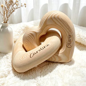 May include: A light-coloured wooden sculpture consisting of two interlocking heart-shaped links. The wood features a visible natural grain pattern. The name Caroline is engraved on the front link, and the name Oliver is engraved on the back link.