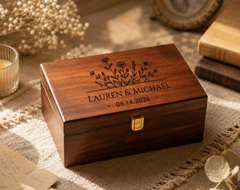 May include: A rectangular dark brown wooden keepsake box with a gold-toned metal latch. The lid features a laser-engraved floral design above the text LAUREN & MICHAEL and the date 06.14.2026, centered in a clean, dark, sans-serif font.