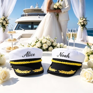 Puede incluir: Dos gorras de capitán blancas y negras con ribete trenzado dorado y detalles de hojas de laurel bordadas en oro en la visera. La gorra izquierda tiene bordado el nombre Alice en letra cursiva negra, y la derecha el nombre Noah en letra negra.
