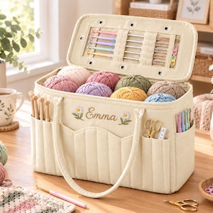 May include: A cream-colored fabric yarn storage bag with the name Emma embroidered in brown script flanked by daisy motifs. The bag features exterior pockets holding wooden hooks and scissors, while the open lid displays various crochet hooks.